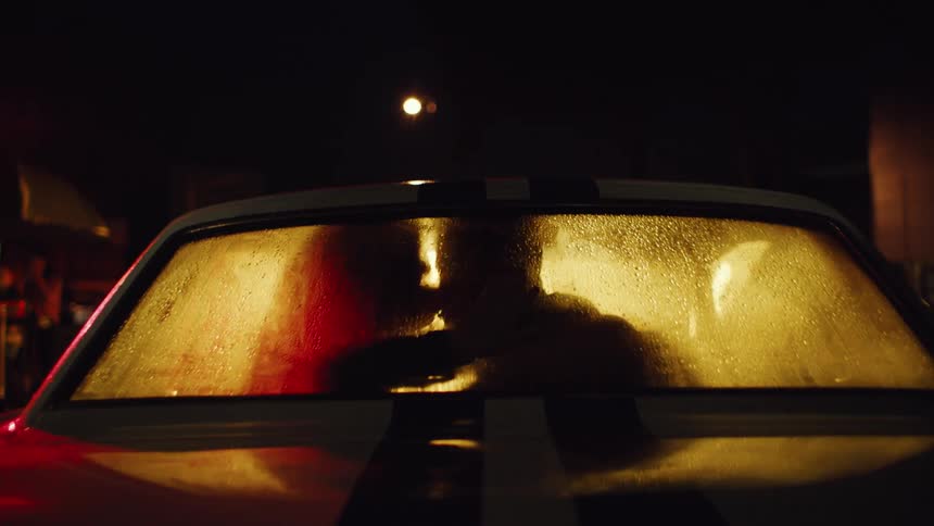A man and a woman kiss in the backseat of a car on a city street at night