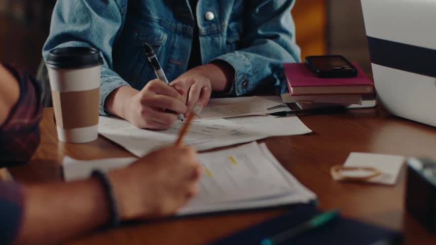 Two young people at a table writing on printed papers with coffee cups notebooks and a phone on the table