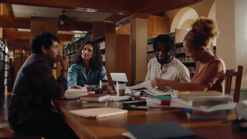 Group of four friends sitting at a table talking and reading books in a library