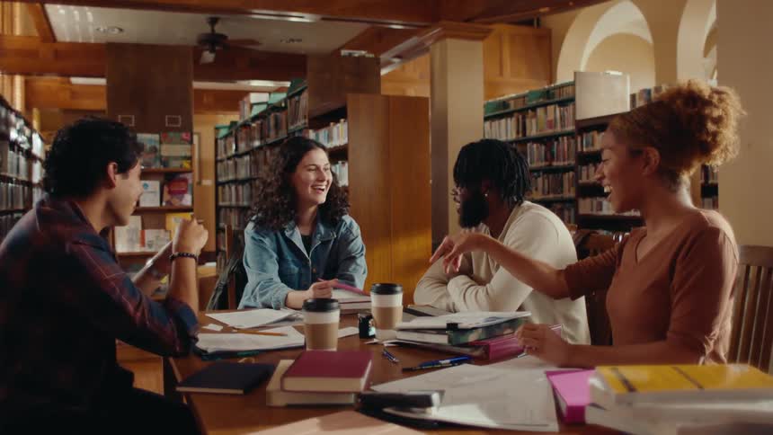 A group of four individuals laughing in a study session at a library table and one throws a paper airplane
