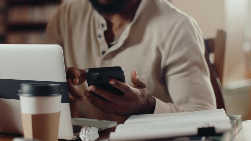A young man is sitting in a library looking straight ahead and talking to someone and then takes out his cell phone and looks to the side and continues talking