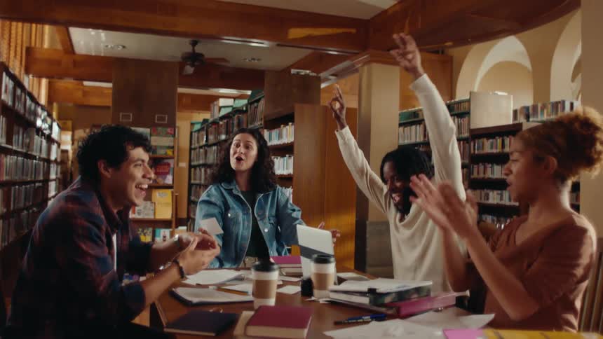 Friends sitting at a library table raising their hands cheering smiling