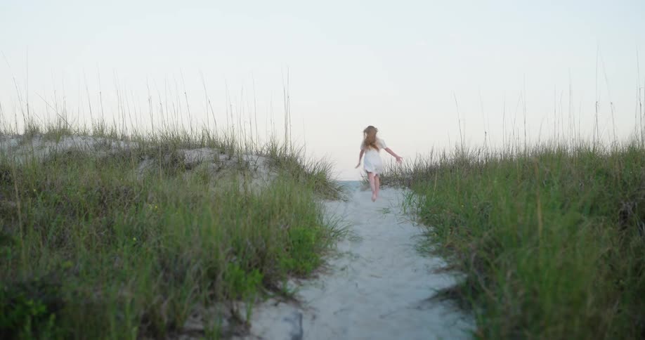 Young female running between grass along a path near the coast kicks up sand