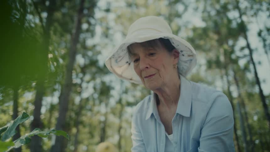 Older woman smiles while picking in her garden near a wooded area