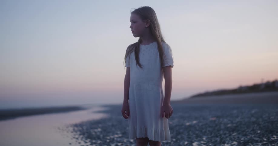 A little girl in a white dress is on the beach looking at the ocean