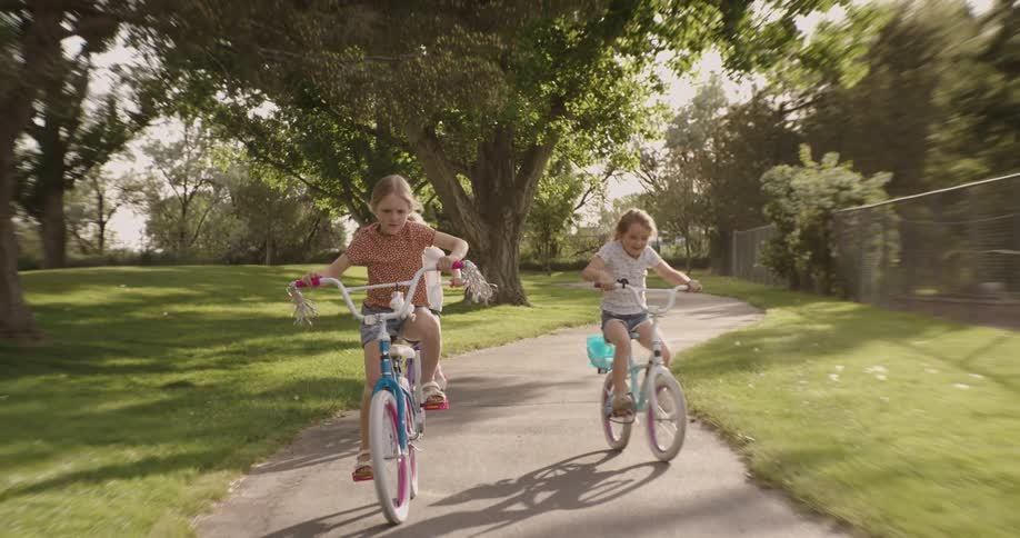 Three little blonde girls are riding bikes on a concrete path next to lawns