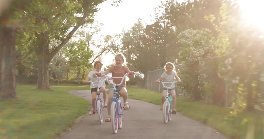 Three girls smile while riding their bicycles along a path through the park on a sunny summer day