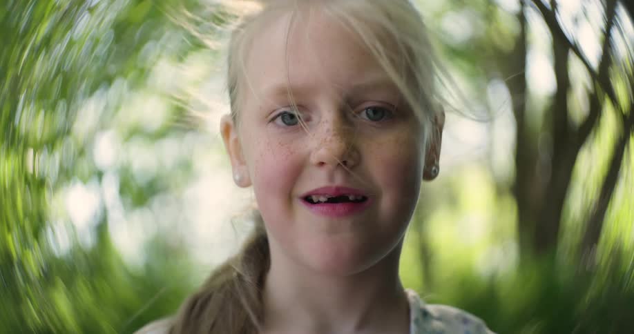 A little blonde girl with missing teeth and freckles smiles with trees in the background