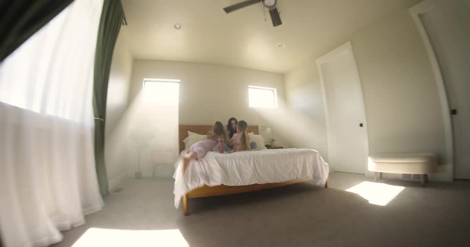A young girl runs and jumps on the bed with her mother and two sisters