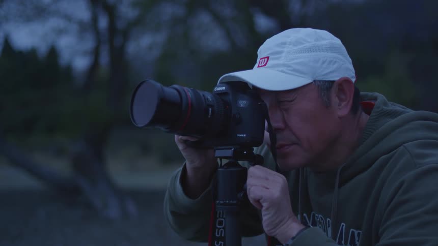 A man in a white cap is fiddling with a professional camera that is standing on a tripod and he is looking through the lens