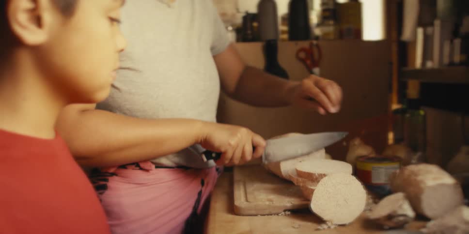 Son accompanying his mother holding a knife and cutting a vegetable on top of a wooden board