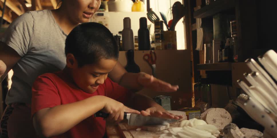 A mother teaches her son how to chop vegetables on a cutting board