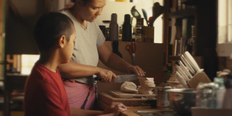 A woman is cutting food into slices and there is a boy next to her who is watching