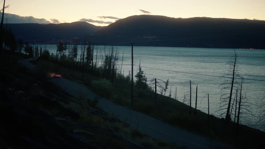 Car driving along a lakeside road at dusk with mountains in the background