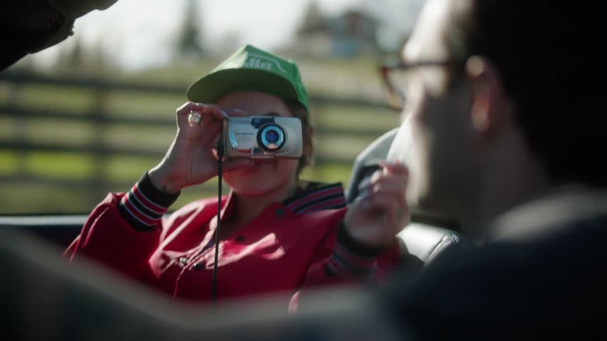 Man driving a convertible car and a woman next to him taking pictures with a camera