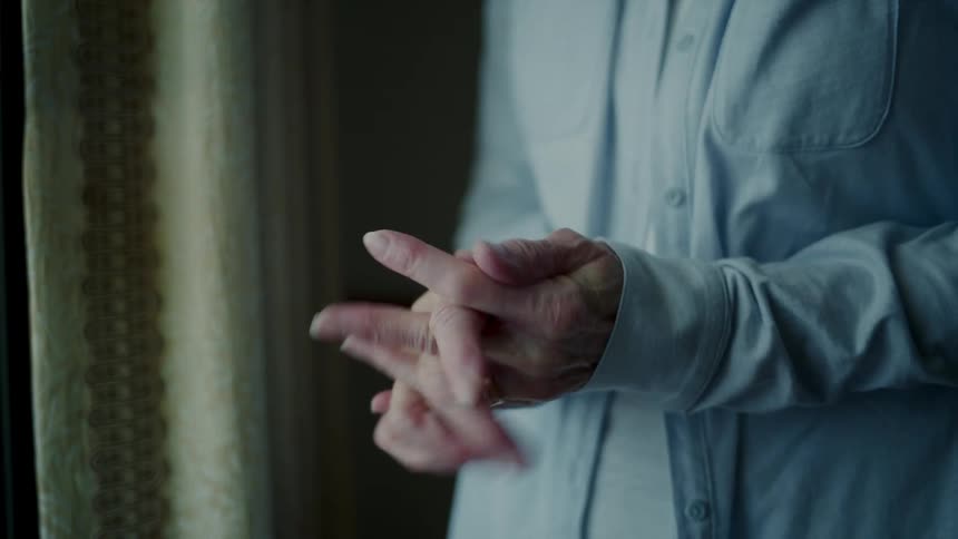 A woman wearing a blue shirt stands inside a house rubbing her hands together nervously
