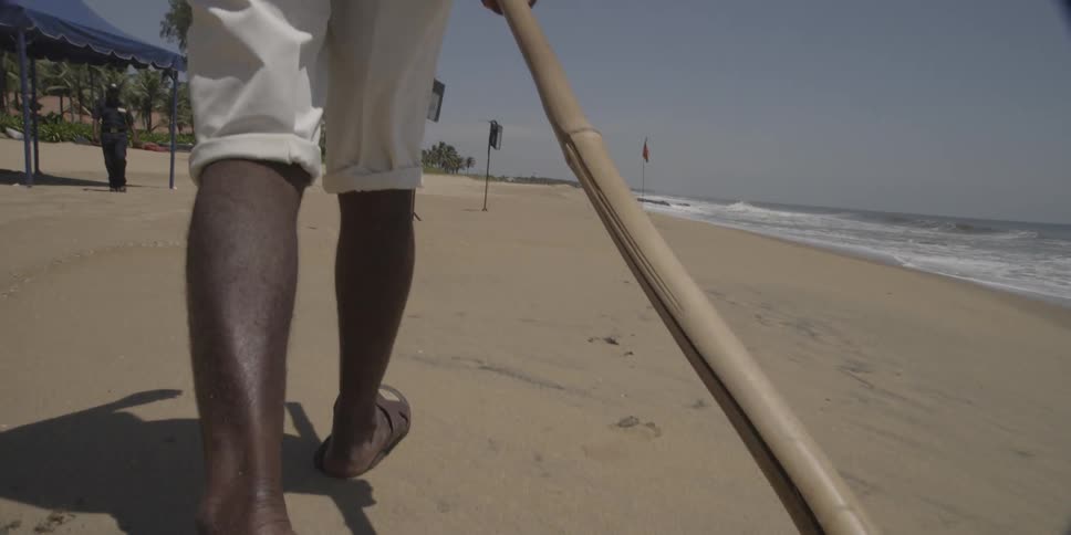 A man is walking on a beach dragging a wooden rake behind him