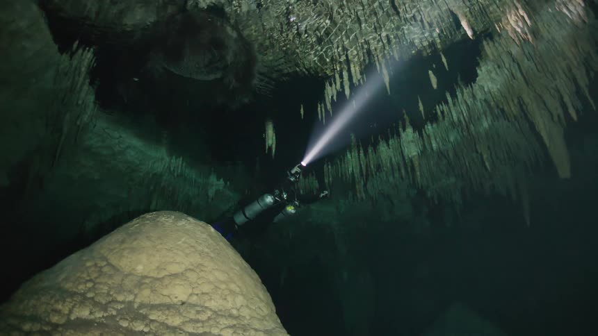 A diver explores an underwater cave using a flashlight to illuminate the stalactites hanging from the ceiling