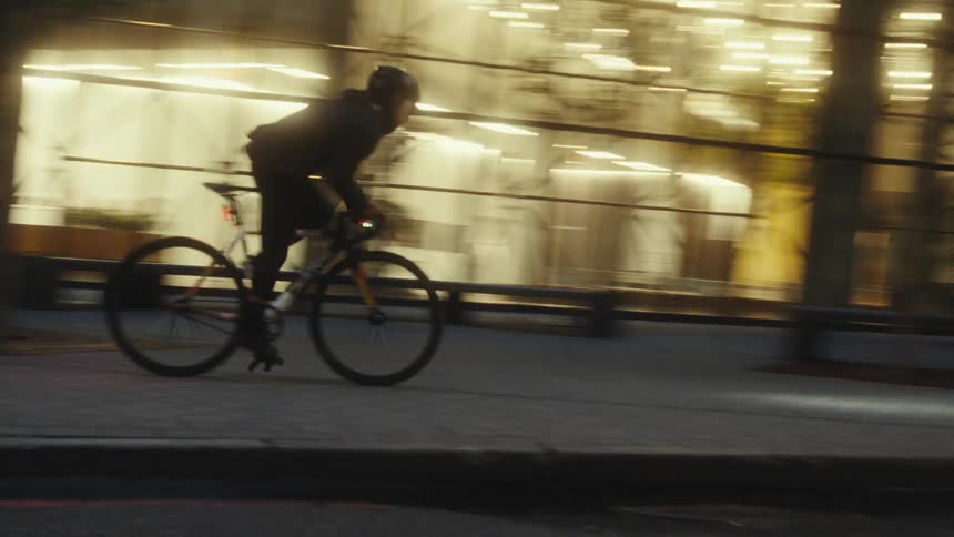 Woman rides a bicycle quickly down a city block past a building with large windows just before dark