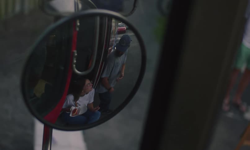 Two people eat food outside a food truck as shown in a rear view mirror