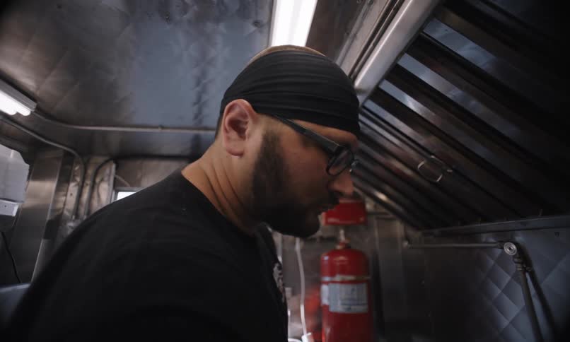A man with glasses and a black bandana on his head is fiddling with something on a counter in front of him