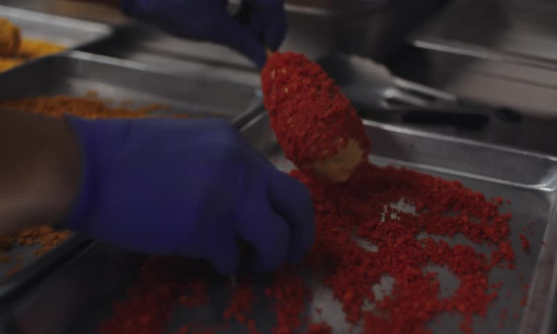 Human hands wearing blue gloves are preparing a food on a stick and spreading red flour around it