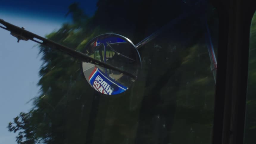 A rearview mirror that is reflecting a blue truck that is driving in a place where many trees pass by
