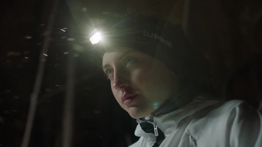 Woman with wearing a headlamp blows into her hands as the snow blows around her