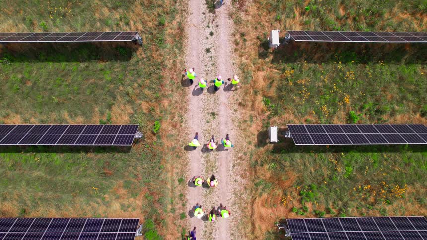 An aerial view of a solar farm with several individuals in high visibility vests walking on a dirt path between rows of solar panels