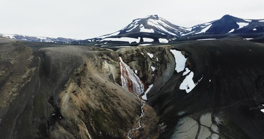 A small waterfall coming out of a hill in a mountainous region with some snow on the ground