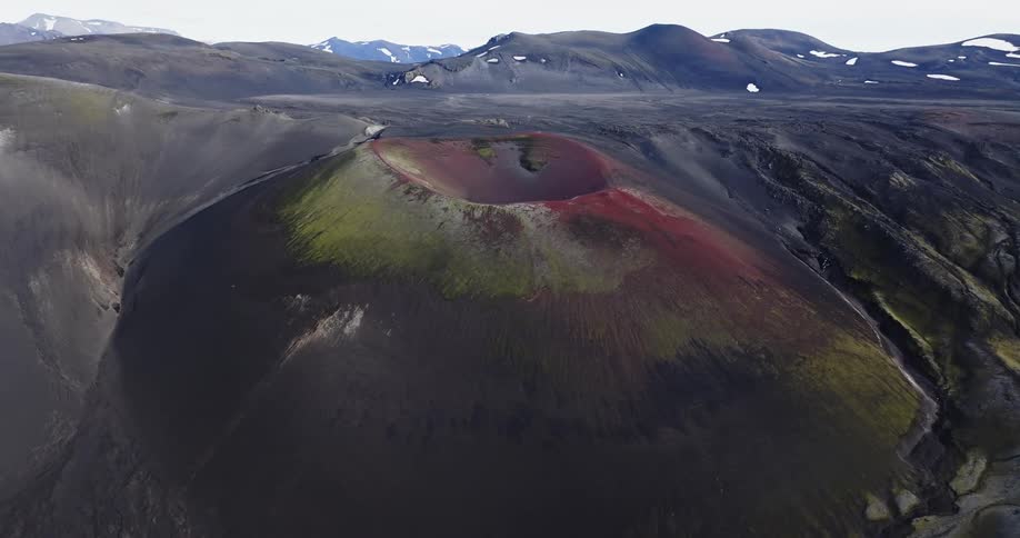 Aerial view of a volcanic crater with a distinct area of red color contrasting with the surrounding dark volcanic soil