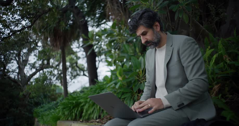 Man types on a laptop while sitting on a wall in a park