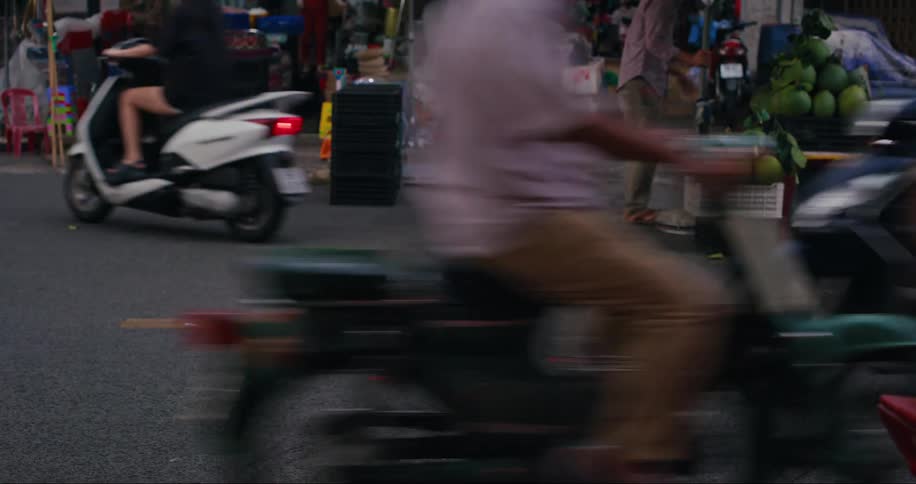 A busy city street with several people riding on mopeds motor scooters and motorcycles in front of a fruit market