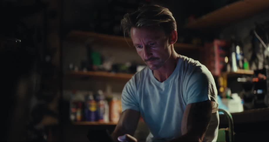 A man sitting in a garage by a motorcycle uses a smartphone to text