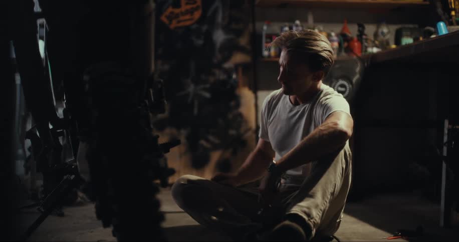 A man sits on the floor in a cluttered garage while he repairs his motorcycle