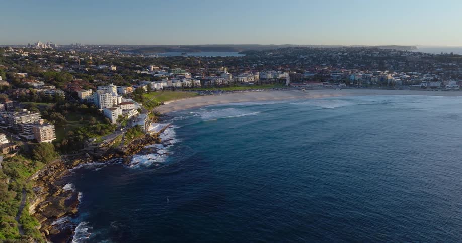 Aerial view of a long beach with light sand surrounded by residential buildings and vegetation with a deep blue sea to the side