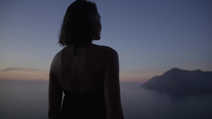 Woman from behind contemplates a serene landscape of mountains and sea during dawn