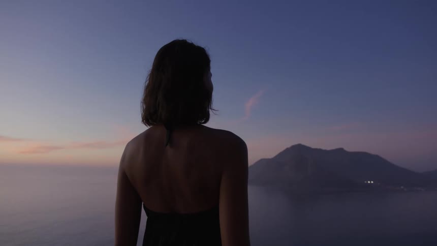 View of a woman from behind enjoying the view by the sea with the silhouette of mountains on the horizon in a purpletoned dawn