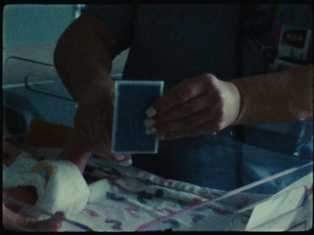 Female nurse presses flat rectangular object to the hand of a baby sleeping in a hospital bassinet