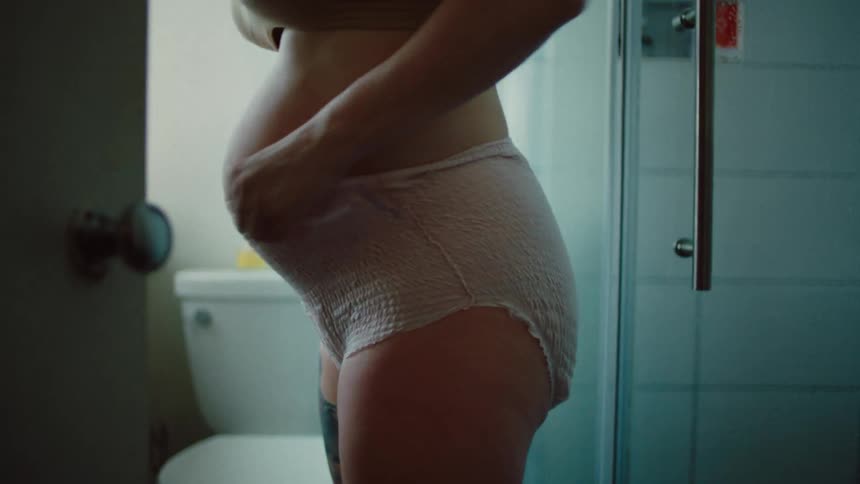 A pregnant woman is putting on shorts and passing them between her belly