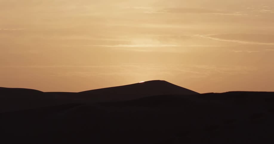 A desert landscape during sunset the sun is partially hidden behind a sand dune casting a warm glow across the horizon
