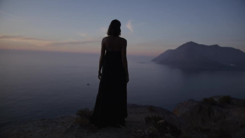 Woman in a long flowy dress walks on a large walk leading to an ocean with mountains in the distance