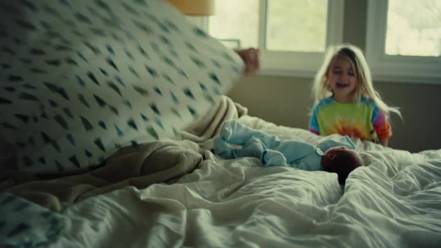 Mother sitting on the bed adjusting the blanket with a newborn baby in front of her and a little girl next to her laughing