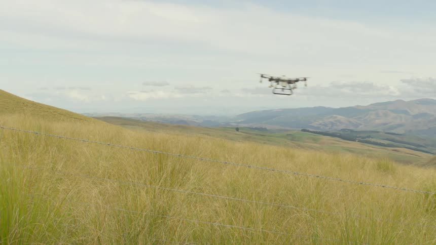 Drone flying over a field of tall grass with a fence in the foreground and hills in the background under a partly cloudy sky