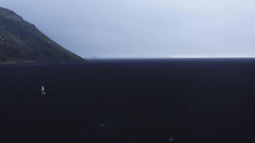 A person wearing all white walks and stumbles over a vast black plain near a mountain