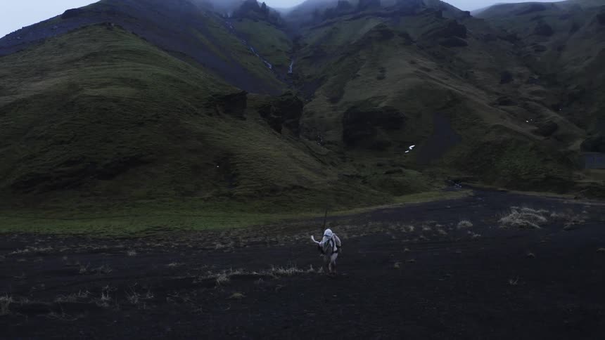 A person walks across a plain near mountains dressed in white robes while holding a staff