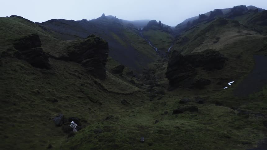 Aerial view of a person wearing a white cape walking through a mountain valley