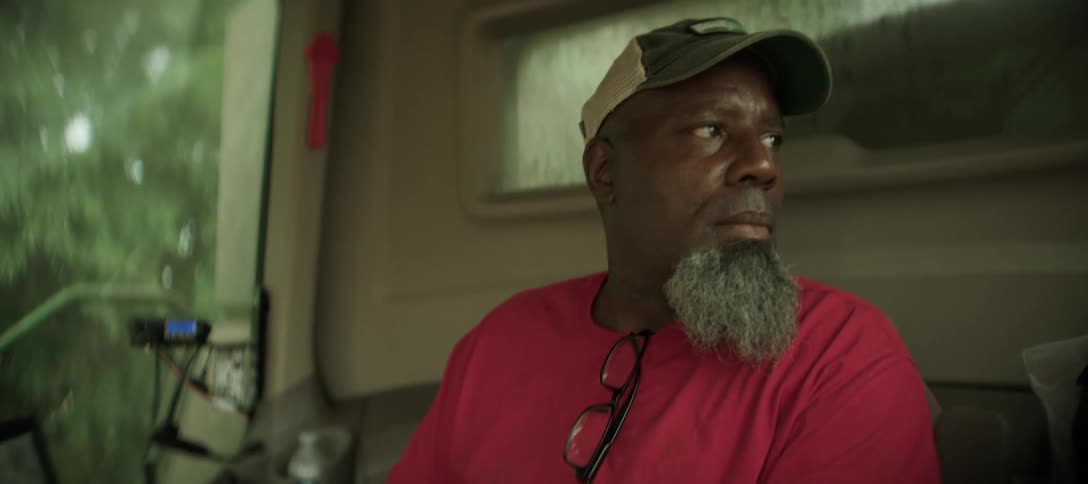 A bearded man wearing a baseball cap and red shirt sits on the porch looking to the right
