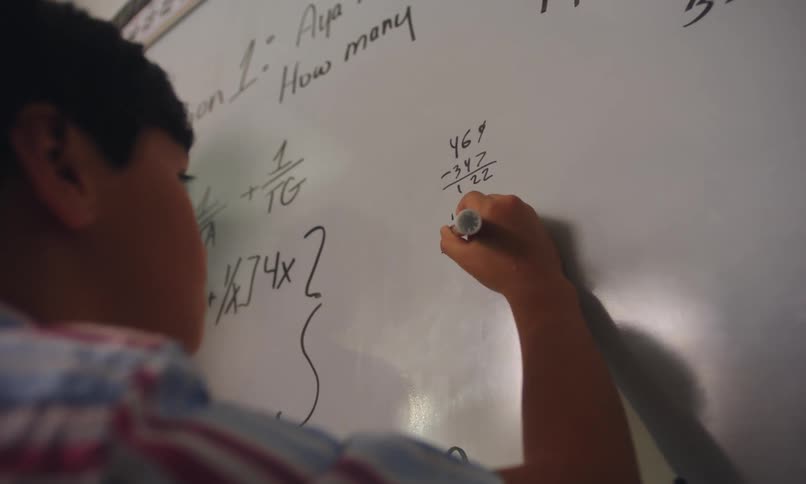 A young boy writes a math problem on a whiteboard while standing next to his teacher