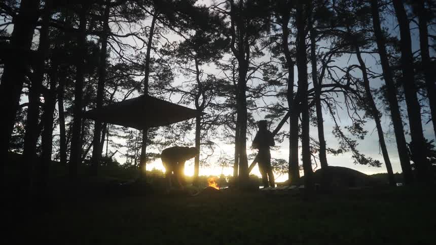 Two people building a fire on a forest with a tarp connected to trees to provide shelter